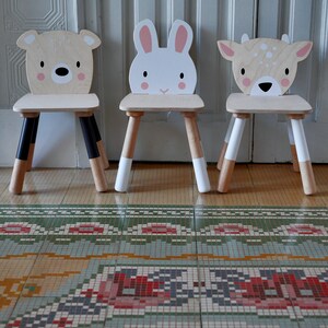 May include: Three wooden children's chairs with animal faces. The chairs are painted in white, brown, and black. The chairs are on a tiled floor with a patterned rug.