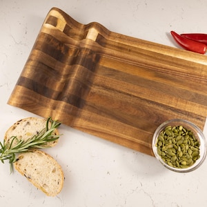 Op de afbeelding: Een houten snijplank met een golvende rand, met een rijke bruine kleur en houtnerf. Twee sneetjes brood met rozemarijn, een kleine glazen kom met groene pompoenpitten en twee rode chilipepers zijn op de plank gerangschikt. De plank is ongeveer 46 cm lang.