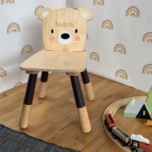 May include: A wooden children's chair with a bear-shaped backrest. The chair is painted brown and has black accents on the legs. The backrest has the name "Teddy" written on it.