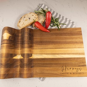 May include: A wooden chopping board with a curved edge, engraved with "Harry's CHEESE & BISCUITS." Slices of bread, red chillies, and herbs are arranged on a striped cloth, suggesting a food preparation theme.