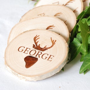 May include: Wooden coasters with a deer head design and personalized names. The coasters are made from natural wood slices and feature engraved text. The word "GEORGE" is visible on the front coaster. Green leaves are in the background.