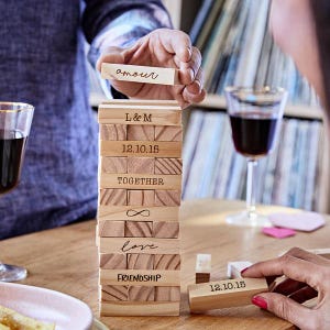 May include: A wooden block stacking game with personalized engravings. The blocks are engraved with words like "amour", "love", and "friendship", and dates. A hand is holding a block that says "amour".