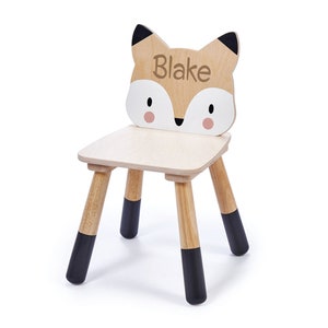 May include: A wooden children's chair with a fox face on the back. The chair is painted brown with black accents and the fox face has a white nose and pink cheeks. The name "Blake" is written on the fox's forehead.