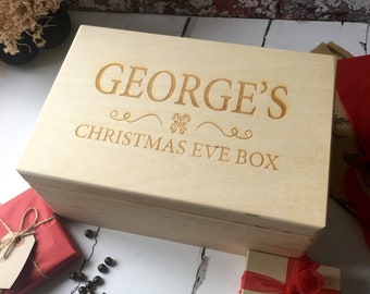 Puede incluir: Una caja de madera con el texto "GEORGE'S CHRISTMAS EVE BOX" grabado en la tapa. La caja es de color marrón claro y tiene un acabado de madera natural.
