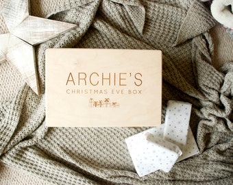 Puede incluir: Una caja de madera con el texto "ARCHIE'S CHRISTMAS EVE BOX" grabado en la parte superior. La caja está sobre una manta de punto beige.