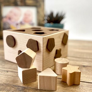 Può includere: Giocattolo di smistamento di forme in legno con finitura naturale. Il cubo ha varie forme ritagliate e include forme in legno come un cerchio, un pentagono, una stella e una casa. Un classico giocattolo educativo per bambini piccoli.