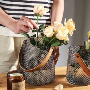 May include: Two clear glass vases with a diamond pattern and brown leather handles. The vases are filled with water and yellow roses.