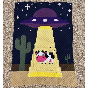Puede incluir: Una manta de ganchillo con un diseño de OVNI con una parte superior morada y un rayo amarillo, levantando una vaca. El fondo es un cielo nocturno azul oscuro con estrellas y cactus. La manta está hecha de hilo.