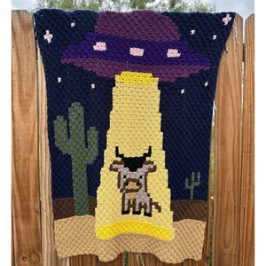 Puede incluir: Manta de ganchillo que representa un OVNI abduciendo a una vaca en un desierto. La manta tiene un cielo nocturno azul oscuro con estrellas, un OVNI morado y rosa, un rayo amarillo y cactus verdes. La vaca es de color canela y negro.