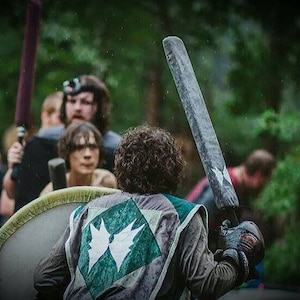 May include: A group of people dressed in medieval garb are participating in a live action role-playing game. One person is holding a large wooden sword, while another person is holding a shield. The scene is set in a wooded area.