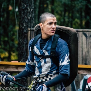 May include: A person wearing a blue and white patterned shirt with a shield on their back, holding a weapon. The shirt has a unique design with white accents. The background includes a wooden fence and trees.