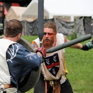 May include: Two people in medieval garb are engaged in a sword fight. One person is wearing a white tunic with a black and white design, and the other is wearing a white tunic with a red and black design. The person on the left is holding a shield, and the person on the right is holding a sword.