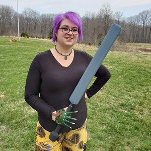 May include: A person with purple hair is holding a grey foam sword with a green and brown leather claw hand grip. The person is wearing a black long-sleeved shirt and yellow and green patterned pants.