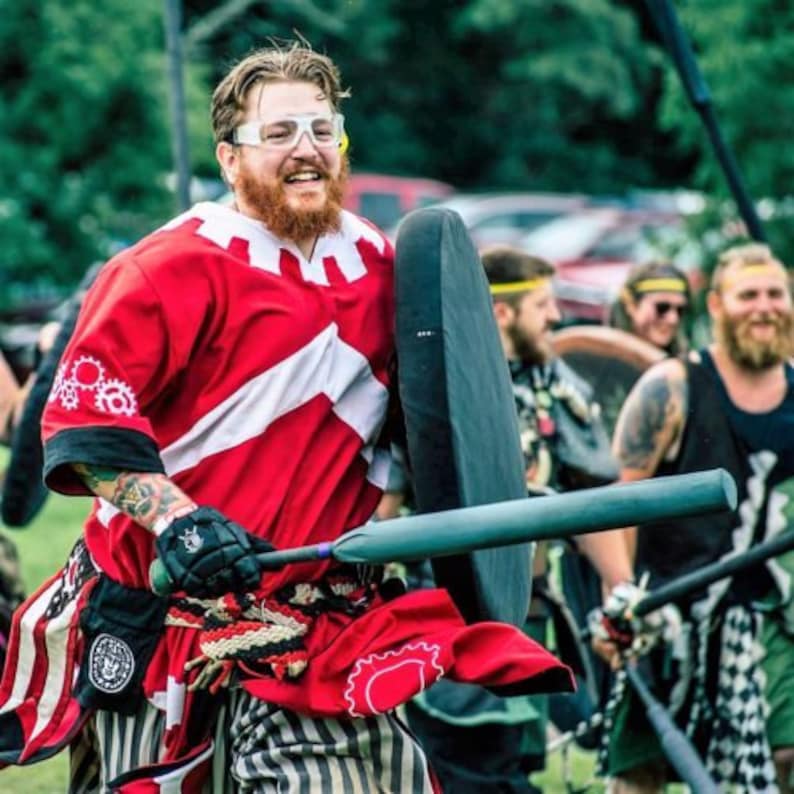 May include: A man wearing a red and white shirt with a black and white striped skirt holds a green and black foam-covered bat and a black shield. He is wearing clear safety glasses and has a beard.