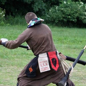 May include: A person wearing a brown tunic with a colorful scarf around their neck is holding a wooden staff. The tunic has a black sash with a red and white patch that says "Ragnarok XXX" and "Red Weapon Champion".
