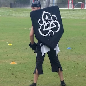 May include: A person wearing black pants and a white shirt is holding a black shield with a white symbol on it. The shield is shaped like a triangle with rounded edges. The symbol is a circle with a triangle inside of it. The person is standing on a grassy field.