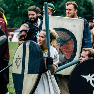 May include: A group of people dressed in medieval garb, holding shields and swords. One person is wearing a white tunic and holding a large, round shield with a black and white design. The shield has a star-shaped symbol in the center.