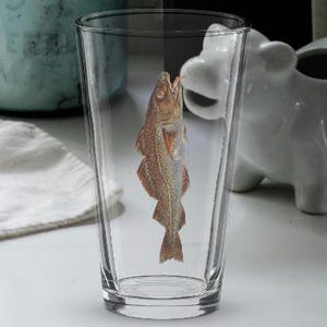 Könnte beinhalten: Ein klares Glas mit einem aufgedruckten Fisch steht auf einer weißen Arbeitsplatte. Eine weiße Sparschwein-Spardose und ein blaues Glas befinden sich im Hintergrund. Ein weißes und gemustertes Handtuch liegt auf der Theke.