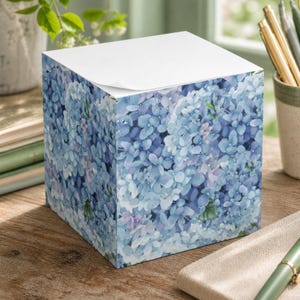May include: A square paper memo cube featuring a repeating pattern of blue and light purple hydrangea flowers on all visible sides. The top surface consists of a stack of plain white square paper sheets with a slight curl at the corner.