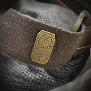 May include: A rectangular brass-toned metal pin featuring an intricate geometric maze-like pattern with recessed lines. The pin is attached to a dark brown textured fabric strap that has visible stitching and a metallic loop component at the top edge.