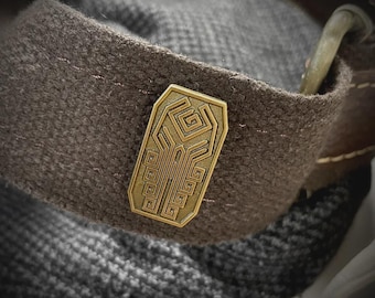 May include: A rectangular brass-toned metal pin featuring an intricate geometric maze-like pattern with recessed lines. The pin is attached to a dark brown textured fabric strap that has visible stitching and a metallic loop component at the top edge.