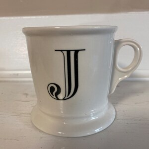 Może przedstawiać: Biały ceramiczny kubek z czarną literą "J" w ozdobnej czcionce. Kubek ma uchwyt i podstawę. Jest przeznaczony do gorących napoj&oacute;w, takich jak kawa lub herbata.