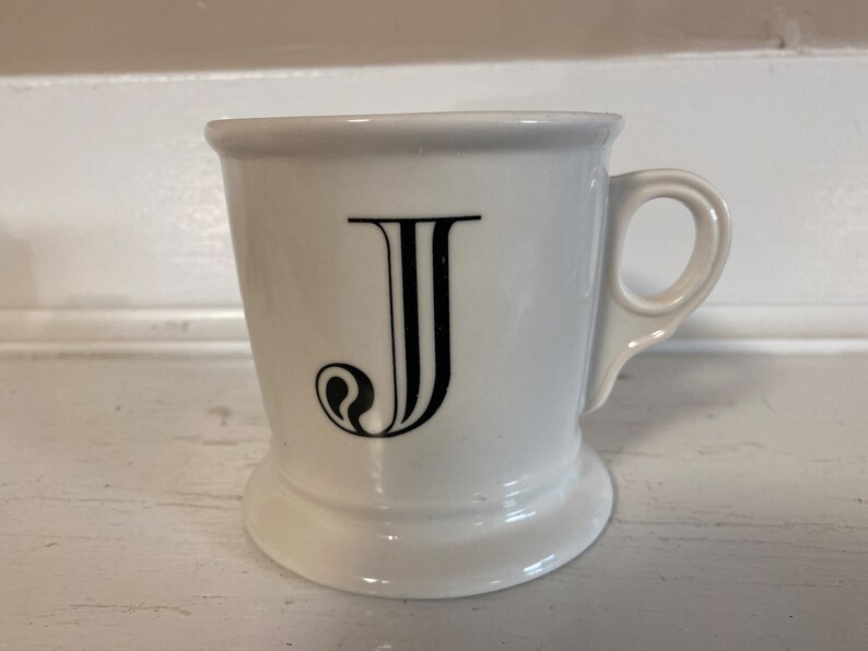 Może przedstawiać: Biały ceramiczny kubek z czarną literą "J" w ozdobnej czcionce. Kubek ma uchwyt i podstawę. Jest przeznaczony do gorących napoj&oacute;w, takich jak kawa lub herbata.