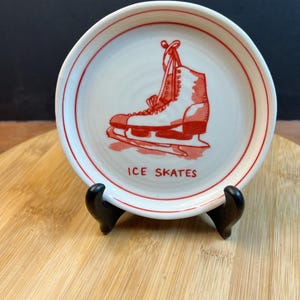 May include: A small, round, white ceramic plate with a red border and a red illustration of an ice skate. The words "ICE SKATES" are printed in red below the image. The plate is displayed on a black stand.