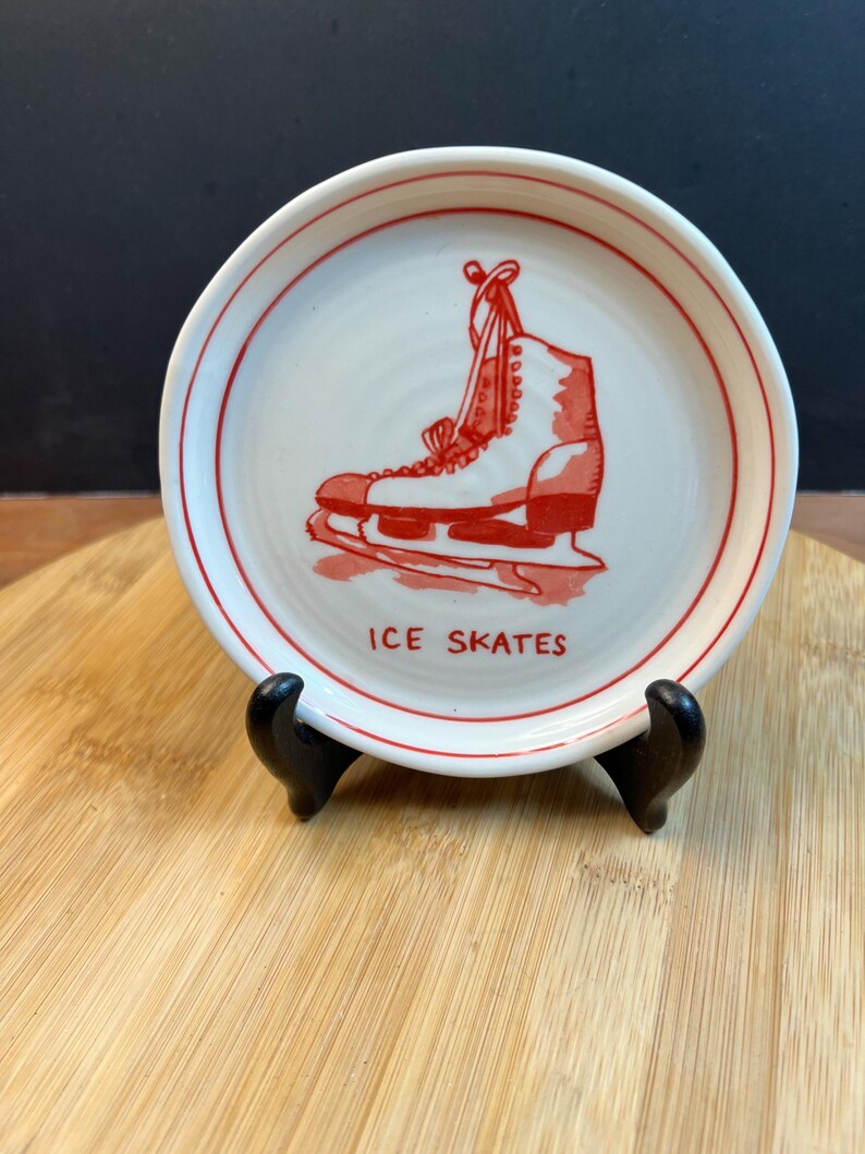 May include: A small, round, white ceramic plate with a red border and a red illustration of an ice skate. The words "ICE SKATES" are printed in red below the image. The plate is displayed on a black stand.