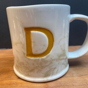 Puede incluir: Taza de cer&aacute;mica blanca con una gran letra "D" dorada en la parte delantera. La taza tiene un patr&oacute;n veteado beige y un asa curvada. Sobre una superficie de madera.
