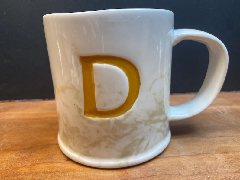 Puede incluir: Taza de cer&aacute;mica blanca con una gran letra "D" dorada en la parte delantera. La taza tiene un patr&oacute;n veteado beige y un asa curvada. Sobre una superficie de madera.