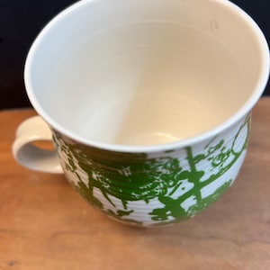 May include: Cream-colored ceramic mug with a green tree design. The mug has a rounded shape with a handle and a wide opening. The green design is a repeating pattern of trees. The mug sits on a wooden surface.