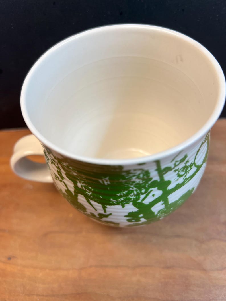 May include: Cream-colored ceramic mug with a green tree design. The mug has a rounded shape with a handle and a wide opening. The green design is a repeating pattern of trees. The mug sits on a wooden surface.