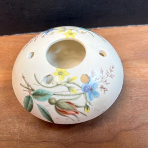 May include: Cream-colored ceramic floral arrangement holder with a central hole and smaller side holes. The holder is decorated with colorful floral designs in yellow, blue, and red, with green leaves and brown stems.