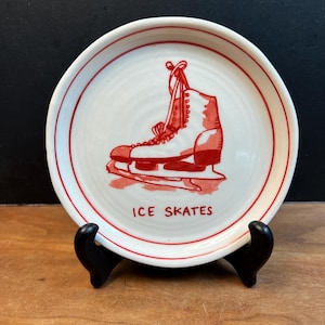 May include: A white ceramic plate with a red ice skate illustration and the words "ICE SKATES". The plate has a red rim and is displayed on a black stand against a dark background.