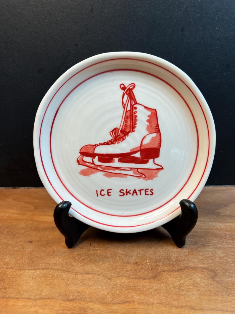 May include: A white ceramic plate with a red ice skate illustration and the words "ICE SKATES". The plate has a red rim and is displayed on a black stand against a dark background.