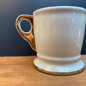 May include: A white ceramic mug with a gold rim and handle. The mug has a wide base and a rounded body.
