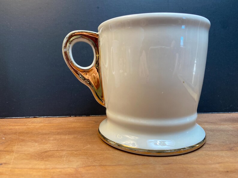 May include: A white ceramic mug with a gold rim and handle. The mug has a wide base and a rounded body.