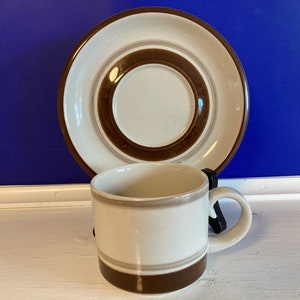 Pode incluir: Uma xícara de café de cerâmica branca com listras marrons e um pires combinando. O pires tem um anel marrom ao redor do centro.