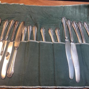 May include: A set of silver cutlery, including knives, forks, and spoons, is arranged in a green fabric roll. The utensils have decorative handles and are displayed against a dark background.