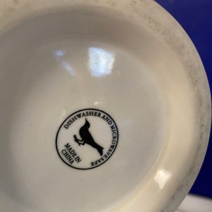 Może przedstawiać: Sp&oacute;d białego ceramicznego kubka z czarną okrągłą etykietą. Na etykiecie widnieje napis "Można myć w zmywarce i kuchence mikrofalowej" oraz "Made in China". Czarna sylwetka ptaka znajduje się na środku etykiety.