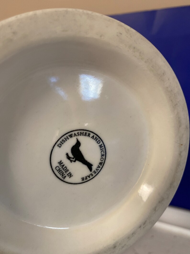 Może przedstawiać: Sp&oacute;d białego ceramicznego kubka z czarną okrągłą etykietą. Na etykiecie widnieje napis "Można myć w zmywarce i kuchence mikrofalowej" oraz "Made in China". Czarna sylwetka ptaka znajduje się na środku etykiety.