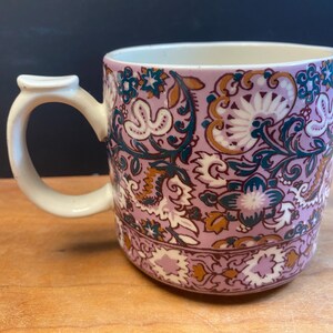 May include: A ceramic mug with a floral pattern in lavender, white, teal, and gold. The mug has a white handle and a cream interior. The design features stylized flowers and leaves, creating a vintage aesthetic. The mug is suitable for tea or coffee.