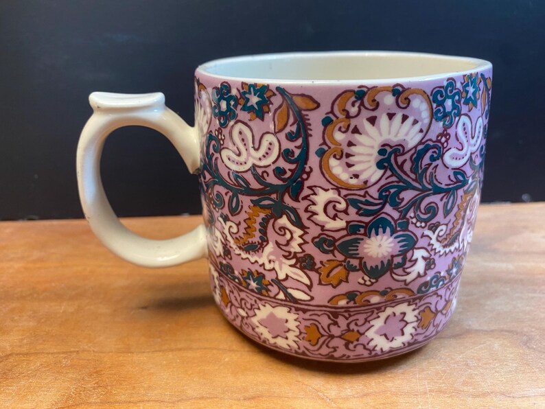 May include: A ceramic mug with a floral pattern in lavender, white, teal, and gold. The mug has a white handle and a cream interior. The design features stylized flowers and leaves, creating a vintage aesthetic. The mug is suitable for tea or coffee.