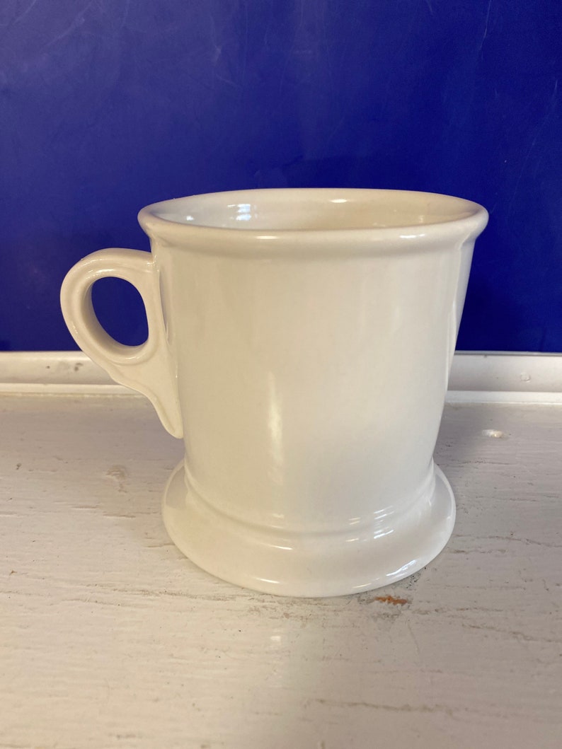 Może przedstawiać: Biały ceramiczny kubek do golenia z uchwytem i szeroką podstawą. Kubek ma prosty, klasyczny design i jest ustawiony na niebieskim tle.