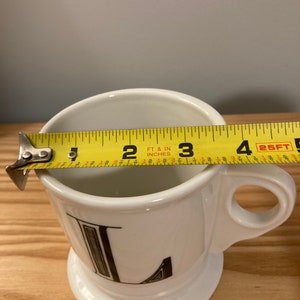 May include: White ceramic mug with a black letter L printed on the front. The mug is 7.6 cm tall and has a handle.