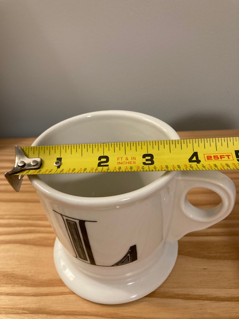 May include: White ceramic mug with a black letter L printed on the front. The mug is 7.6 cm tall and has a handle.