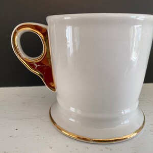 May include: White ceramic shaving mug with a gold rim and a gold-trimmed handle. The handle has a red accent.