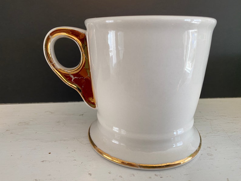 May include: White ceramic shaving mug with a gold rim and a gold-trimmed handle. The handle has a red accent.
