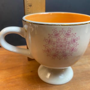 K&ouml;nnte beinhalten: Cremefarbene Teetasse mit wei&szlig;em Henkel und Sockel. Der Innenraum ist orange und der Rand ist goldfarben verziert. Ein rosa Blumendesign verziert die Au&szlig;enseite. Eine dekorative Teetasse zum Servieren von Tee oder Kaffee.